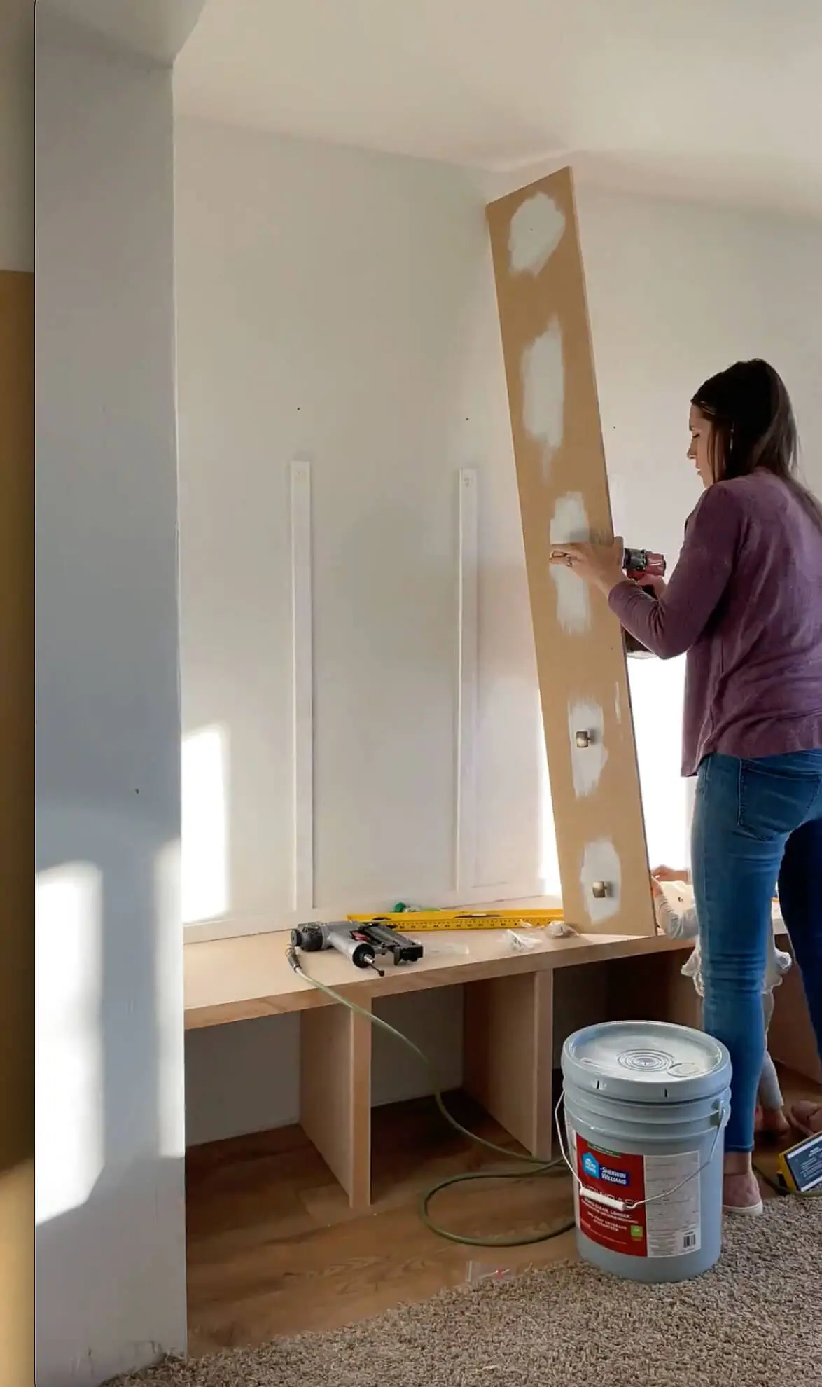 A woman installing a wooden board with pre-filled holes onto a built-in storage bench wall. Various tools, including a nail gun and measuring tape, are scattered on the bench as she continues the DIY home improvement project.